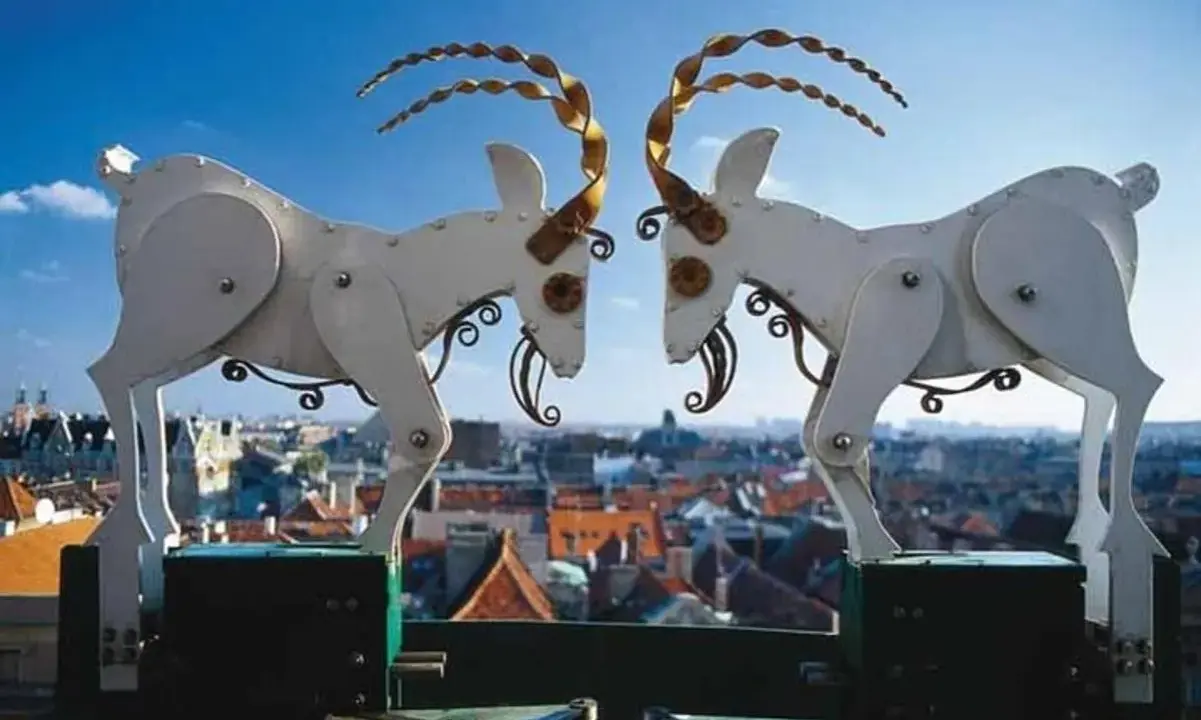 Mechaniczne koziołki poznańskie na tle miasta. Dwa białe, metalowe koziołki z kręconymi rogami stykają się głowami.
