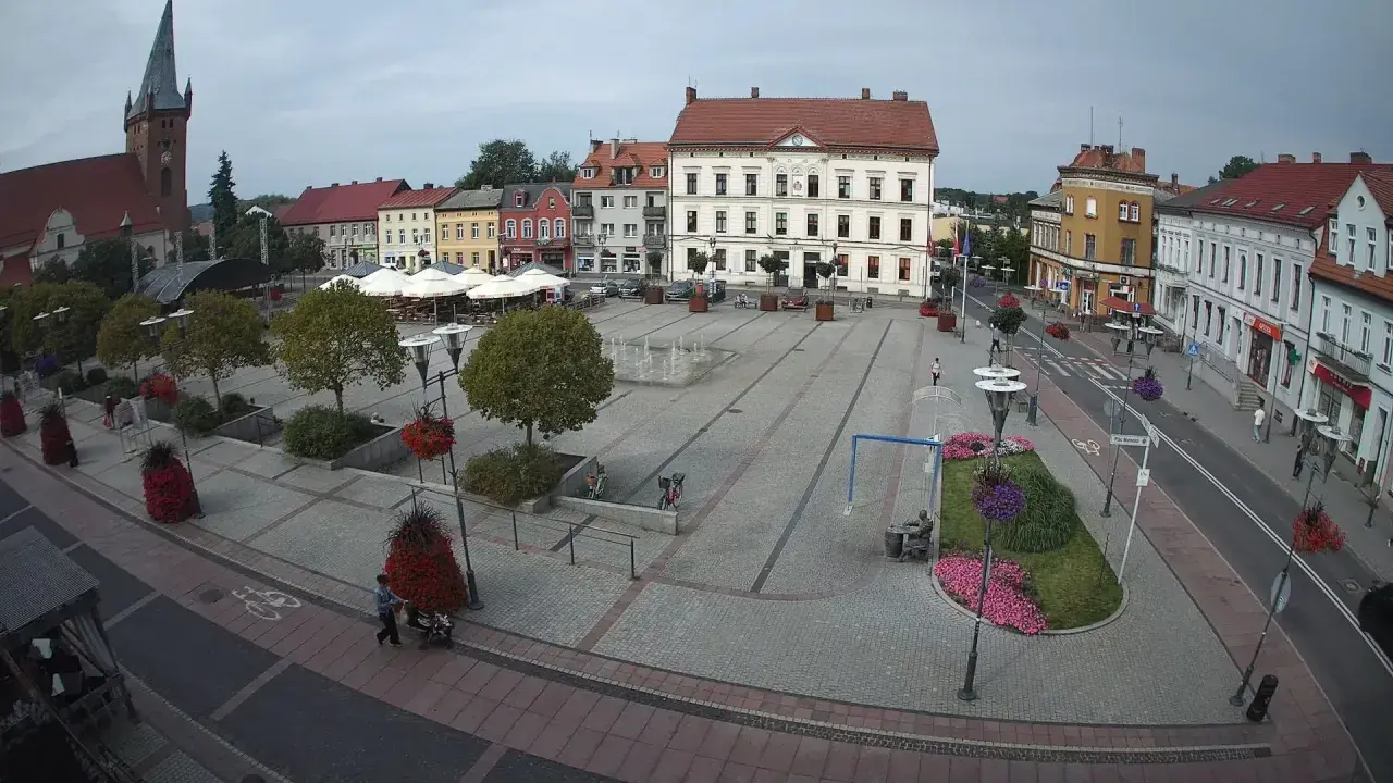 Rynek w Czarnkowie z charakterystycznym kościołem po lewej stronie. Plac tętni życiem, widać ludzi, fontanny i kawiarniane stoliki.