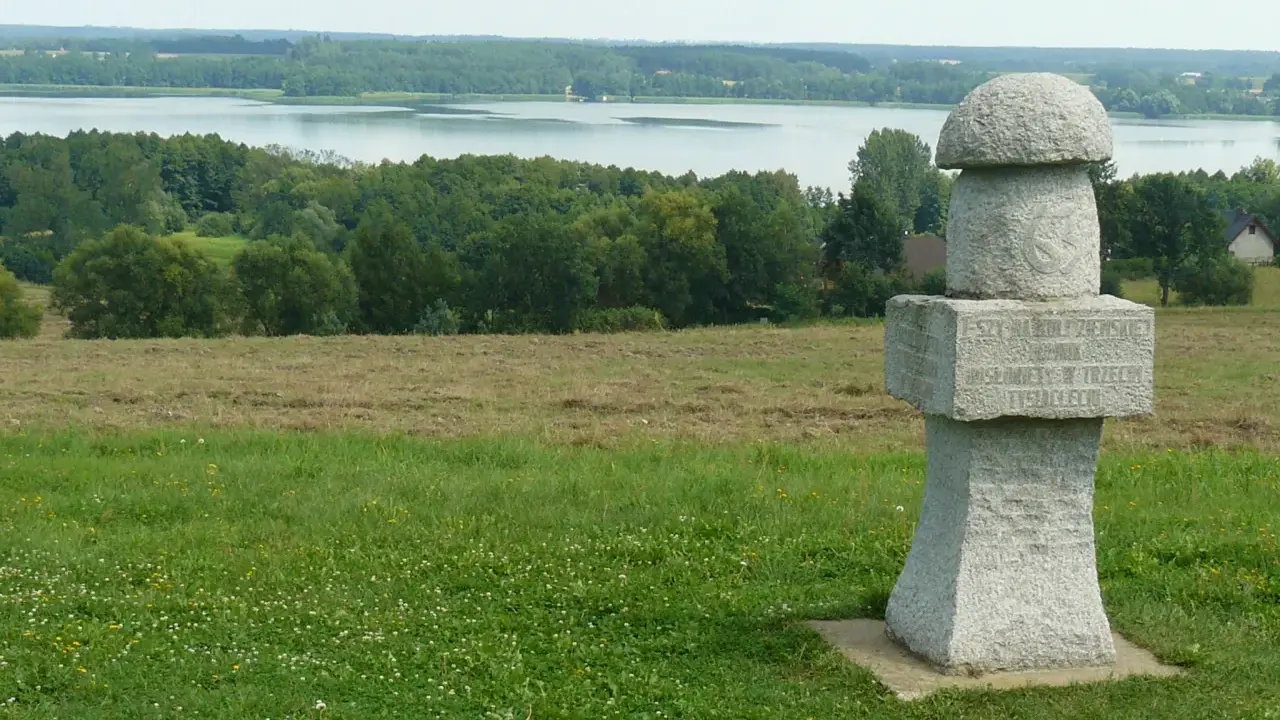 Kamienny, wielki grzyb z inskrypcją stoi na wzgórzu. W tle jezioro i las.