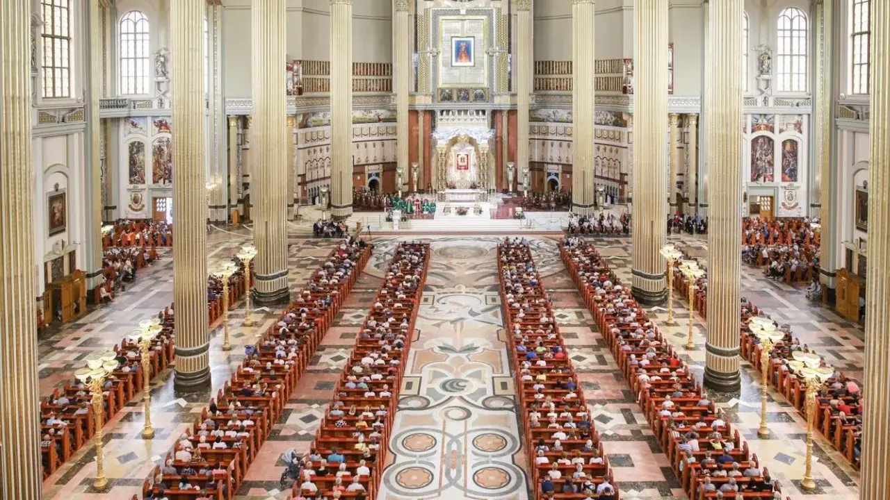 Wielka bazylika w Licheniu Nowym wypełniona wiernymi. Długie ławy zapełnione ludźmi, majestatyczne kolumny i ołtarz w tle.