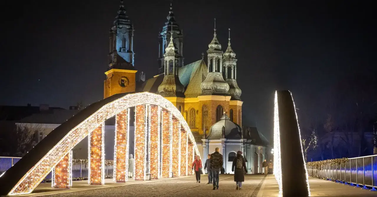 Most Jordana nocą, ozdobiony tysiącami światełek, z katedrą w tle. Spacerujący ludzie podziwiają iluminacje.