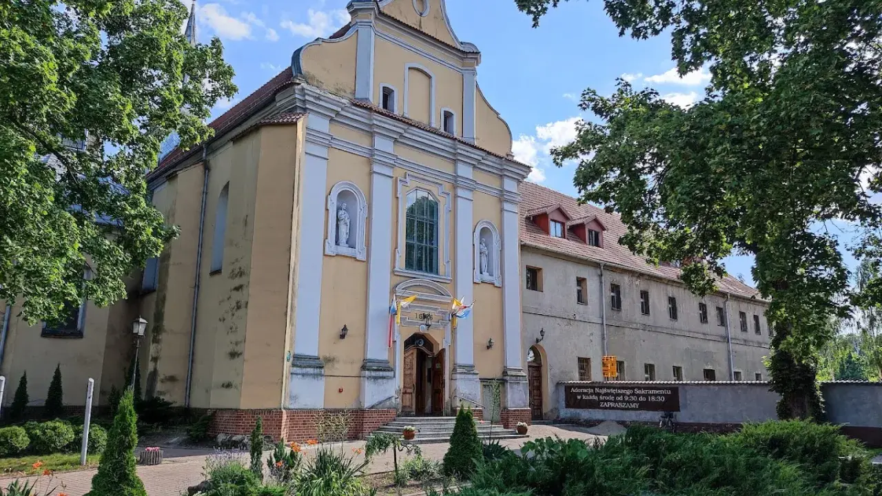 Żółty kościół w Szamotułach z białymi zdobieniami, otoczony zielenią. Obok tablica informująca o adoracji.