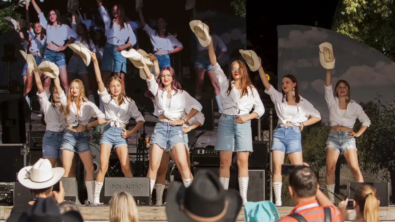 Dziewczęcy zespół country z Wolsztyna w białych koszulach i jeansowych szortach tańczy na scenie, unosząc kapelusze.