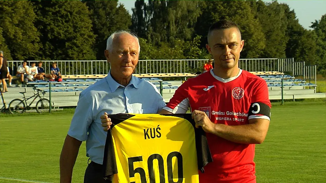 Dwóch mężczyzn na boisku, jeden w niebieskiej koszuli, drugi w czerwonej, trzymają żółtą koszulkę z napisem "KUŚ 500". To ważne wydarzenia Chodzież.