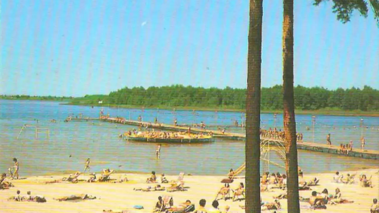 Słoneczny dzień nad jeziorem Niedzięgiel. Ludzie odpoczywają na plaży i pływają, a na wodzie unosi się platforma rekreacyjna.