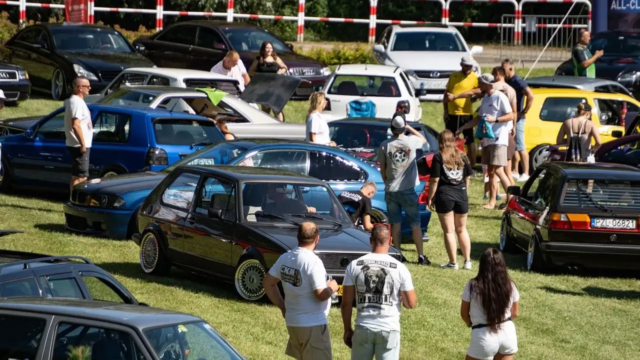 Zlot fanów motoryzacji w Łobżenicy. Wiele samochodów, ludzie oglądają auta, atmosfera piknikowa.