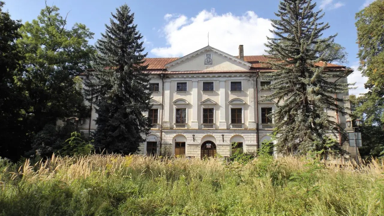 Dawny dwór otoczony przez wysokie trawy i drzewa, w tym sosny i dęby.