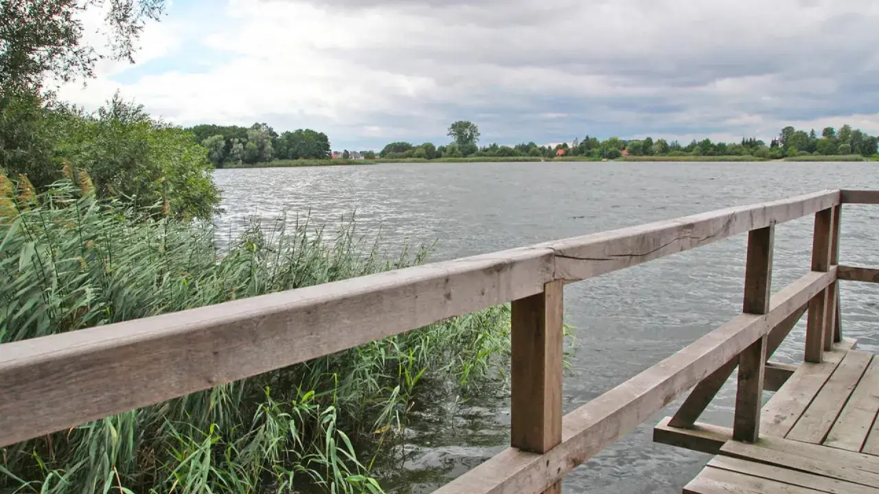 Drewniana kładka nad spokojnym jeziorem Lednica, otoczona trzcinami i zielenią.