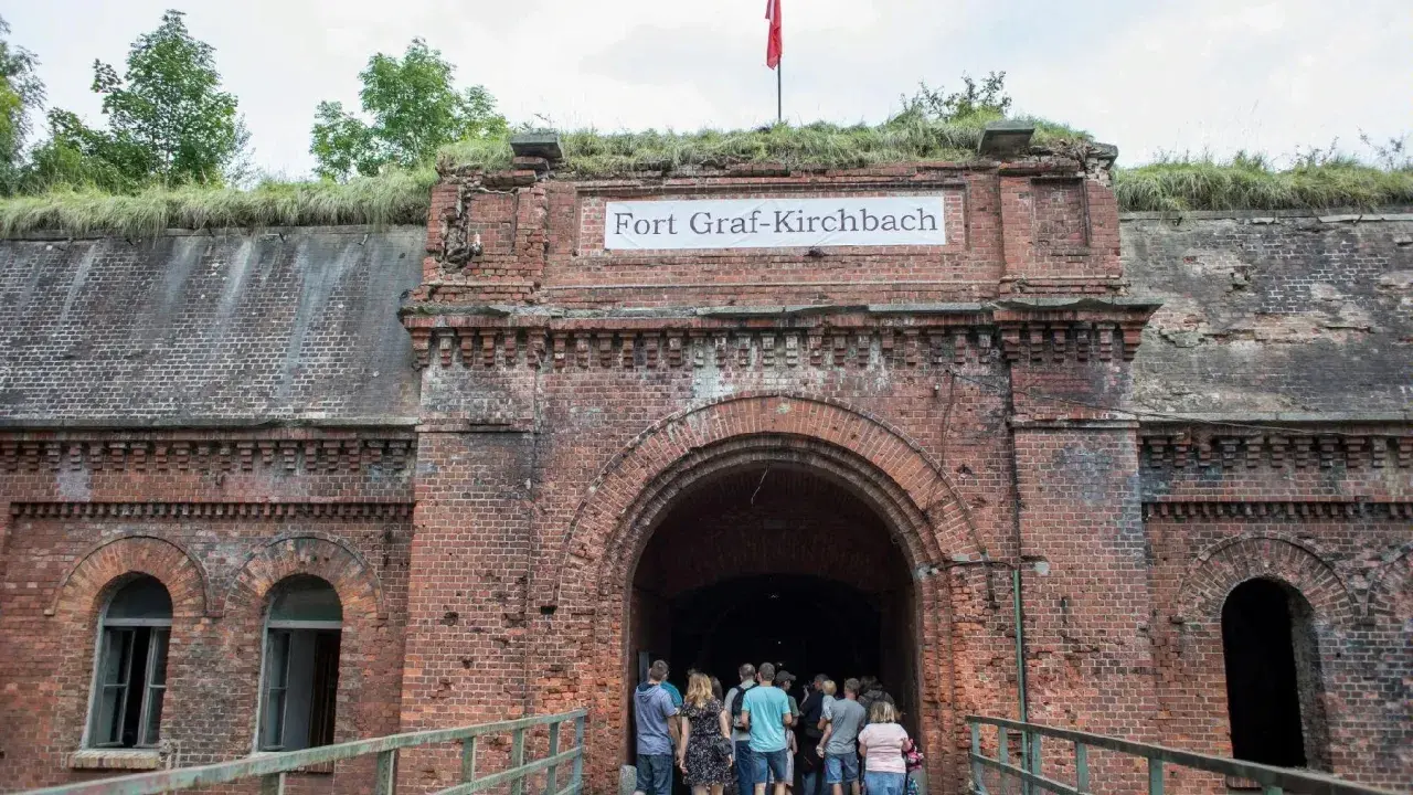 Wejście do Fortu Graf-Kirchbach, historycznej twierdzy w Poznaniu. Grupa turystów wchodzi do ciemnego tunelu.