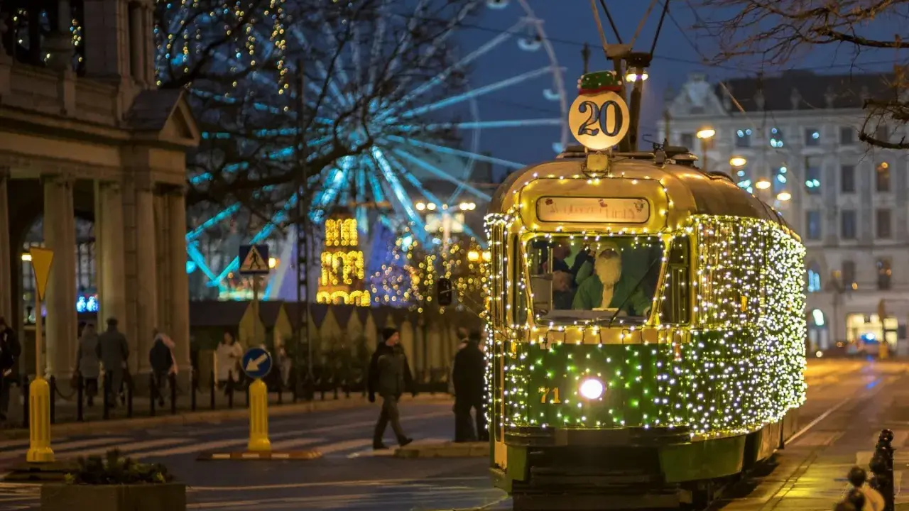 Świąteczny tramwaj po poznańsku, ozdobiony tysiącami lampek, mknie przez miasto. W tle widać diabelski młyn i iluminacje.