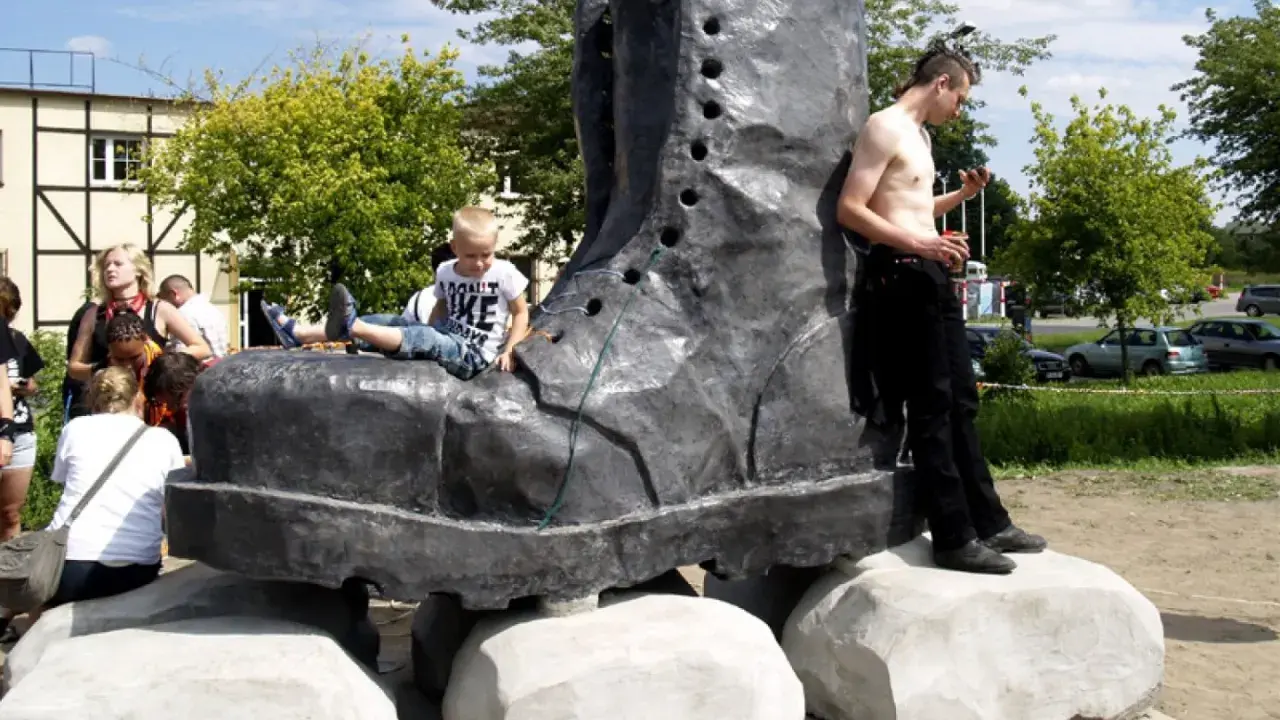 Pomnik glana w kształcie gigantycznego buta, na którym siedzi chłopiec. Obok stoi mężczyzna.