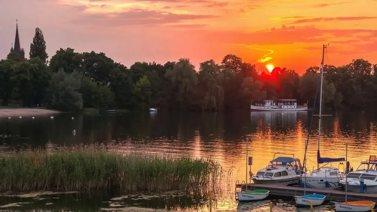 Zachód słońca nad jeziorem z piaszczystą plażą w Polsce. Łodzie zacumowane przy pomoście, na wodzie pływają łabędzie.