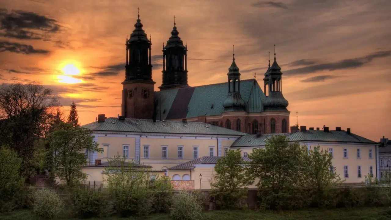 Zachód słońca nad katedrą, najstarszym kościołem w Polsce. Monumentalna budowla z cegły i zielonym dachem, otoczona drzewami.