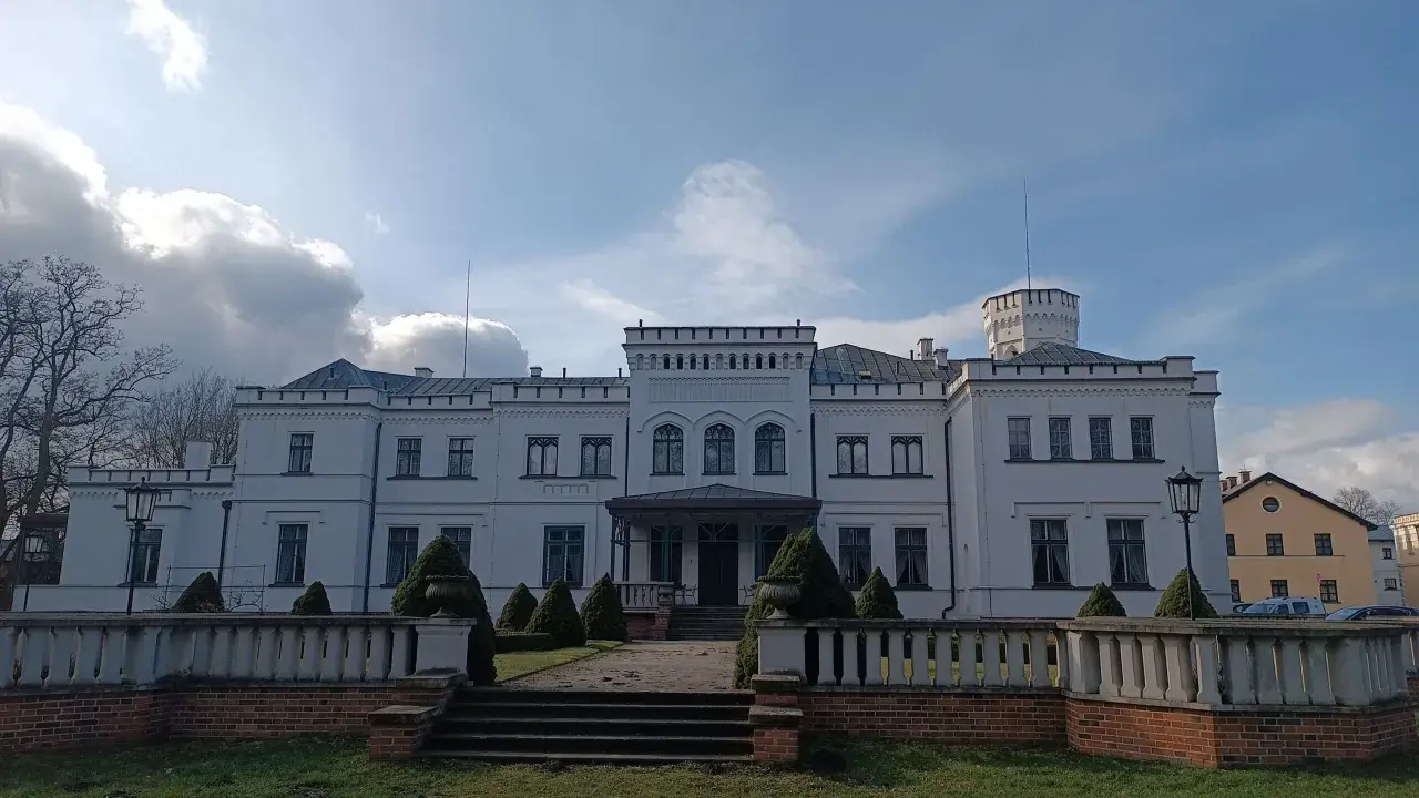 Biały pałac w Będlewie z wieżyczką i ozdobnym murem. Słońce przebija się przez chmury, tworząc malowniczy krajobraz.