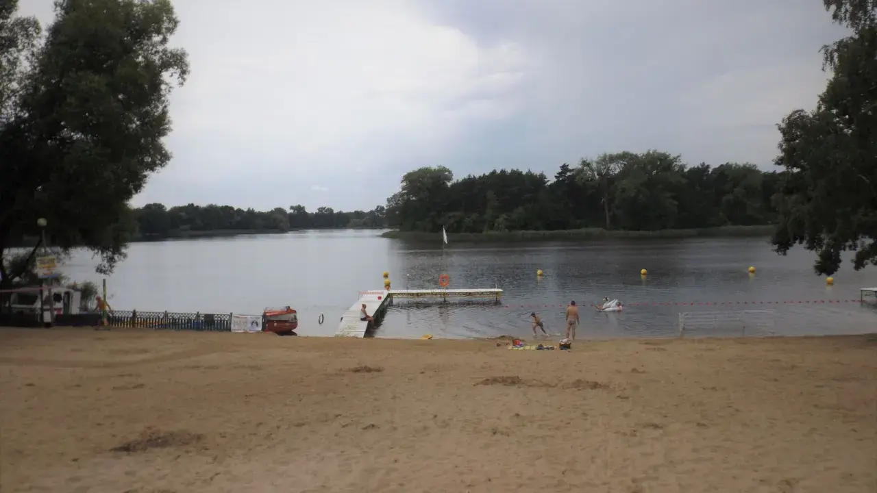 Plaża nad jeziorem koło Gniezna. Ludzie odpoczywają, niektórzy kąpią się. W tle las i spokojna woda.