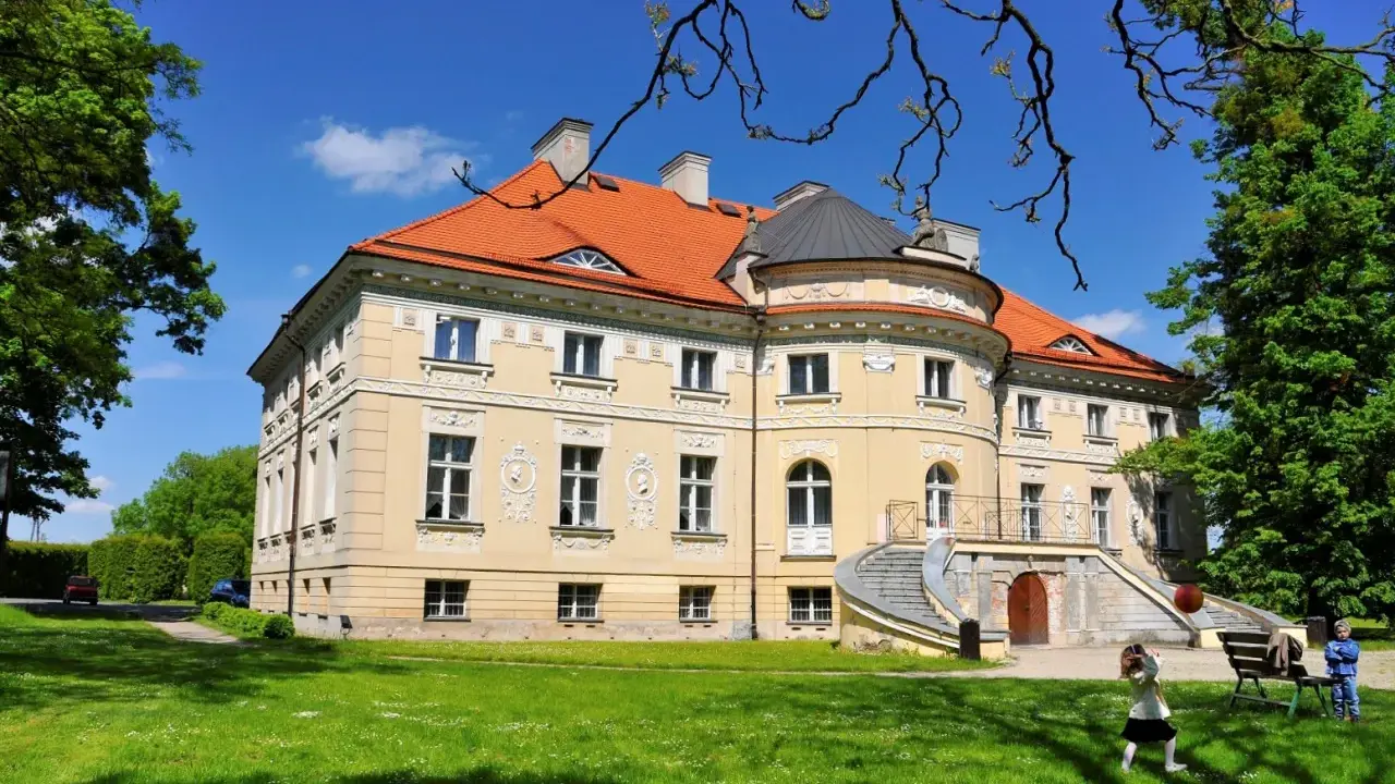 Pałac w Lewkowie, żółty, z pomarańczowym dachem, otoczony zielenią. Dzieci bawią się na trawniku przed budynkiem.