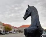 Czarny, metalowy pomnik konia w Koninie, z widokiem na zabytkowe kamienice i samochody.