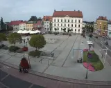 Rynek w Czarnkowie z charakterystycznym kościołem po lewej stronie. Plac tętni życiem, widać ludzi, fontanny i kawiarniane stoliki.