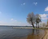Spokojne jezioro w Zbąszyniu, z piaszczystą plażą i pomostem. Drzewa bez liści tworzą malowniczy krajobraz.