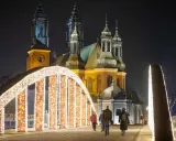 Most Jordana nocą, ozdobiony tysiącami światełek, z katedrą w tle. Spacerujący ludzie podziwiają iluminacje.