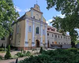 Żółty kościół w Szamotułach z białymi zdobieniami, otoczony zielenią. Obok tablica informująca o adoracji.