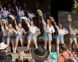 Dziewczęcy zespół country z Wolsztyna w białych koszulach i jeansowych szortach tańczy na scenie, unosząc kapelusze.