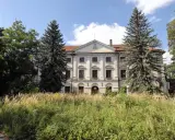 Dawny dwór otoczony przez wysokie trawy i drzewa, w tym sosny i dęby.