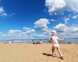 Dziewczynka w różowym kapelusiku bawi się na plaży przy zaporze Jeziorsko. W tle widać ludzi odpoczywających pod parasolem i spokojną taflę wody.