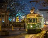 Świąteczny tramwaj po poznańsku, ozdobiony tysiącami lampek, mknie przez miasto. W tle widać diabelski młyn i iluminacje.