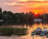 Zachód słońca nad jeziorem z piaszczystą plażą w Polsce. Łodzie zacumowane przy pomoście, na wodzie pływają łabędzie.