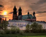 Zachód słońca nad katedrą, najstarszym kościołem w Polsce. Monumentalna budowla z cegły i zielonym dachem, otoczona drzewami.