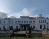 Biały pałac w Będlewie z wieżyczką i ozdobnym murem. Słońce przebija się przez chmury, tworząc malowniczy krajobraz.