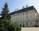 Jasny pałac w ciążeniu, otoczony zielenią, z charakterystycznym dachem i kominami.
