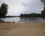 Plaża nad jeziorem koło Gniezna. Ludzie odpoczywają, niektórzy kąpią się. W tle las i spokojna woda.