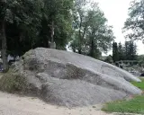Ogromny głaz narzutowy, na którym stoi krzyż. W tle cmentarz i drzewa.