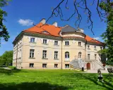 Pałac w Lewkowie, żółty, z pomarańczowym dachem, otoczony zielenią. Dzieci bawią się na trawniku przed budynkiem.
