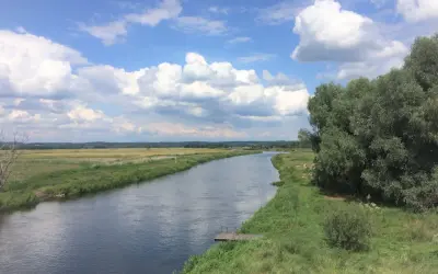 Szeroka rzeka płynie przez zielone łąki i lasy. Na brzegu widać drewniany pomost. To piękny krajobraz Puszczy Noteckiej.
