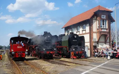 Parada parowozów z parą unoszącą się w powietrzu. Trzy lokomotywy stoją na torach obok ceglanego budynku stacji, a tłum widzów obserwuje wydarzenie.