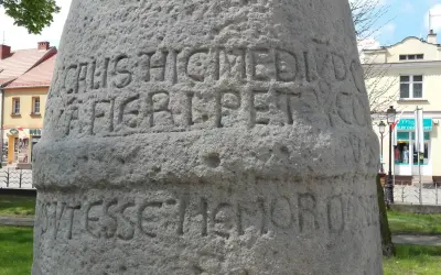 Kamienny słup milowy z wyrytymi łacińskimi napisami, stojący na tle budynków i zieleni.