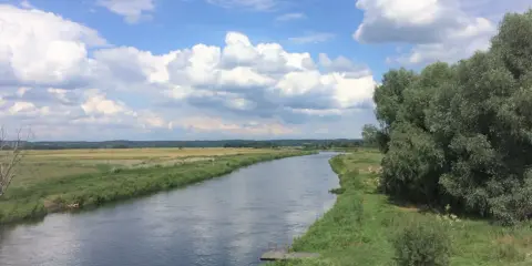 Szeroka rzeka płynie przez zielone łąki i lasy. Na brzegu widać drewniany pomost. To piękny krajobraz Puszczy Noteckiej.