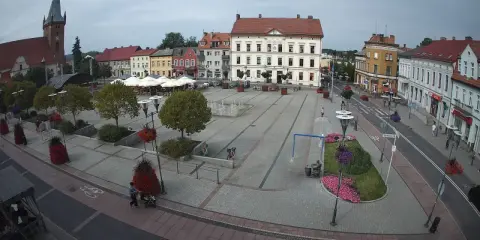 Rynek w Czarnkowie z charakterystycznym kościołem po lewej stronie. Plac tętni życiem, widać ludzi, fontanny i kawiarniane stoliki.