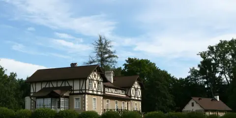 Elegancka zabudowa szachulcowa z ciemnym dachem i białymi ścianami, otoczona zielenią, pod błękitnym niebem.