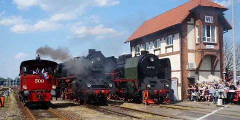 Parada parowozów z parą unoszącą się w powietrzu. Trzy lokomotywy stoją na torach obok ceglanego budynku stacji, a tłum widzów obserwuje wydarzenie.