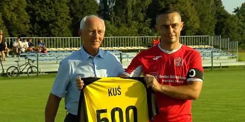 Dwóch mężczyzn na boisku, jeden w niebieskiej koszuli, drugi w czerwonej, trzymają żółtą koszulkę z napisem "KUŚ 500". To ważne wydarzenia Chodzież.