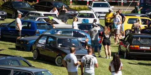 Zlot fanów motoryzacji w Łobżenicy. Wiele samochodów, ludzie oglądają auta, atmosfera piknikowa.
