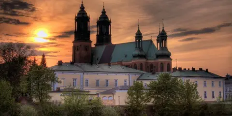 Zachód słońca nad katedrą, najstarszym kościołem w Polsce. Monumentalna budowla z cegły i zielonym dachem, otoczona drzewami.