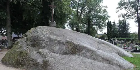 Ogromny głaz narzutowy, na którym stoi krzyż. W tle cmentarz i drzewa.