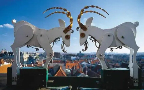 Mechaniczne koziołki poznańskie na tle miasta. Dwa białe, metalowe koziołki z kręconymi rogami stykają się głowami.