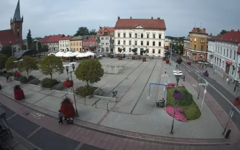 Rynek w Czarnkowie z charakterystycznym kościołem po lewej stronie. Plac tętni życiem, widać ludzi, fontanny i kawiarniane stoliki.