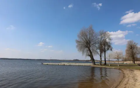 Spokojne jezioro w Zbąszyniu, z piaszczystą plażą i pomostem. Drzewa bez liści tworzą malowniczy krajobraz.